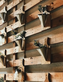 A wall display features a series of wooden pallets with metal cone-shaped planters attached to them. These planters contain green and yellow plants with delicate leaves. Additionally, mason jars with black metal handles are affixed to the wall, adding a rustic decorative element.