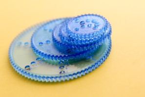 A collection of translucent blue plastic gears of various sizes are stacked on a smooth yellow surface. The gears have evenly spaced holes and intricate teeth along their edges, providing an impression of precision and design.