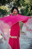 A person is outdoors wearing a bright pink traditional dress adorned with intricate embroidery. They hold a translucent dupatta with floral designs, which appears to be gently flowing. The person is accessorized with ornate jewelry that includes a large necklace, earrings, and a headpiece. Lush green foliage is visible in the background.