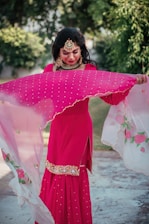 A person is outdoors wearing a bright pink traditional dress adorned with intricate embroidery. They hold a translucent dupatta with floral designs, which appears to be gently flowing. The person is accessorized with ornate jewelry that includes a large necklace, earrings, and a headpiece. Lush green foliage is visible in the background.