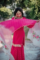 A person is outdoors wearing a bright pink traditional dress adorned with intricate embroidery. They hold a translucent dupatta with floral designs, which appears to be gently flowing. The person is accessorized with ornate jewelry that includes a large necklace, earrings, and a headpiece. Lush green foliage is visible in the background.