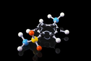 A molecular model composed of various colored spheres connected by rods, representing atoms and bonds. The model is placed against a black background, highlighting the vibrant colors of the spheres including blue, yellow, orange, and white.