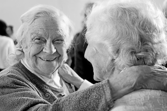 Two elderly people are sharing a joyful moment, with one person facing the camera and smiling warmly while the other is seen from behind, with an arm around the first person's shoulder.