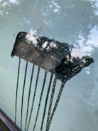 A foggy car windshield with a clear patch where a squeegee or similar tool has wiped away condensation. Water droplets are visible on the cleared area, and tree leaves are reflected in the glass.