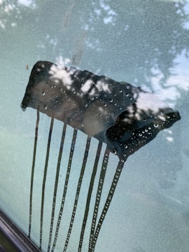 A foggy car windshield with a clear patch where a squeegee or similar tool has wiped away condensation. Water droplets are visible on the cleared area, and tree leaves are reflected in the glass.