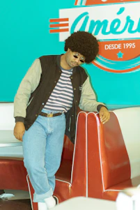 A person with an afro hairstyle and sunglasses leans against a red diner booth. They wear a striped shirt, blue jeans, a black and gray jacket, and white socks with black shoes. In the background, there is a retro-style turquoise wall with a sign that reads 'América DESDE 1995' in white and red.