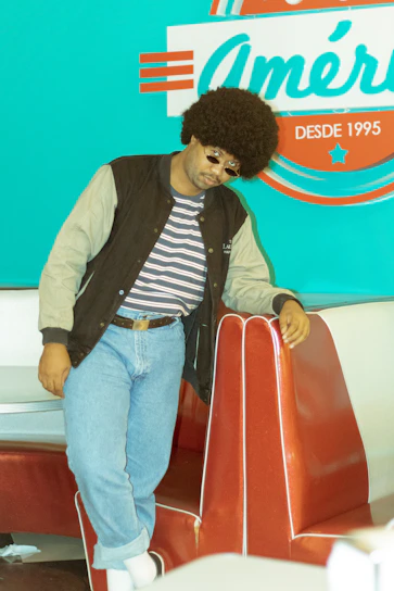 A person with an afro hairstyle and sunglasses leans against a red diner booth. They wear a striped shirt, blue jeans, a black and gray jacket, and white socks with black shoes. In the background, there is a retro-style turquoise wall with a sign that reads 'América DESDE 1995' in white and red.