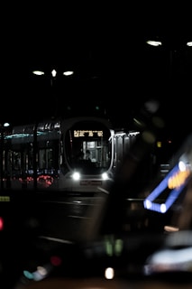 A tram is illuminated in the darkness, with its lights on, casting reflections on the shiny surfaces around. The setting appears to be urban with minimal street lighting visible in the background.