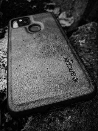 A smartphone with a rugged, textured case lies on a surface covered with rocks. The name 'Ranger' is visible on the case, which has a circular opening for the fingerprint sensor and a camera cut-out on the upper left. The image is captured in black and white, adding a gritty and raw texture to the composition.