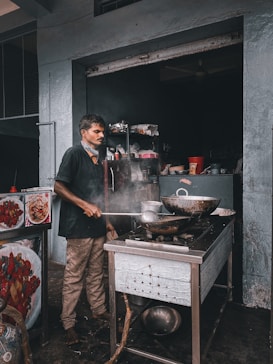 A person cooking in a small kitchen, using a large cooking stove with a wok on top. There is steam rising from the stove as they stir with a long ladle. The kitchen appears rustic and industrial, with shelves holding various containers and cooking utensils. An assortment of items, including a red cup and other kitchen tools, are visible in the background.