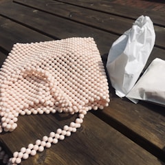 A bead bag with a handle lies on a wooden surface next to two white paper packages. The beads have a light pinkish color, and the wooden table is weathered with a rustic appearance.