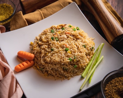 A neatly arranged plate of fried rice garnished with sliced carrots and green vegetables. The rice appears to contain peas, corn, and small pieces of chicken, served on a square white plate. Nearby, there are cooking spices and a wooden pepper grinder on a rustic wooden table.