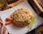 A neatly arranged plate of fried rice garnished with sliced carrots and green vegetables. The rice appears to contain peas, corn, and small pieces of chicken, served on a square white plate. Nearby, there are cooking spices and a wooden pepper grinder on a rustic wooden table.