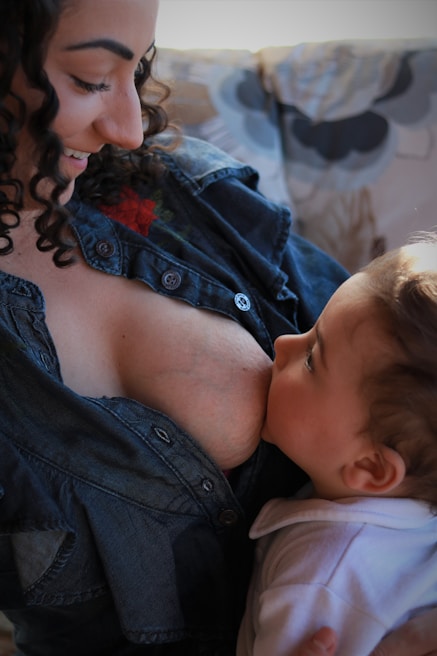 A tender moment between a mother and her baby as she breastfeeds. The mother is wearing a denim shirt and is smiling as she gazes at the baby, who is wrapped in white clothing and looking up at her.