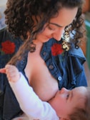 A woman with curly hair is breastfeeding a baby. She is wearing a denim shirt with red floral embroidery. The woman looks down at the baby with a gentle expression. The baby is reaching up and touching her hair.