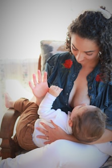 A woman is sitting in a chair breastfeeding a baby. She has curly hair tied back with a scrunchie and is wearing a dark blue top with floral embroidery. The baby is dressed in a white top and brown pants.