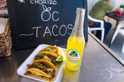 A close-up of a bottle of Jarritos pineapple soda placed on a metal table alongside a dish containing several tacos garnished with cilantro and onion, with a lime wedge. In the background, a chalkboard displays the words 'Chorizo Tacos' with a decorative drawing, and a person is seated at a table.
