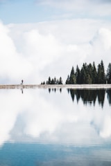 A calm lake reflecting soft clouds with a person walking along the shore.