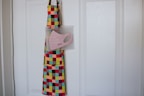 A colorful apron with a checkered pattern in red, green, yellow, and blue hangs on a white door. Attached to the apron is a clear plastic pouch containing a pink face mask, labeled 'Atoms Everyday Face Mask'.