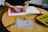 A person is drawing on a stencil while seated at a round wooden table. Various colored templates are stacked, and colored pencils are scattered nearby. On the table lies a transparent pouch labeled 'Atoms Everyday Face Mask'.