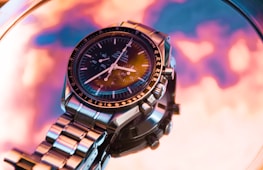 A detailed and elegant wristwatch with a stainless steel band and a black dial, featuring multiple dials and markers. The background is an abstract blend of pink, orange, and purple tones, creating a vibrant and modern aesthetic.