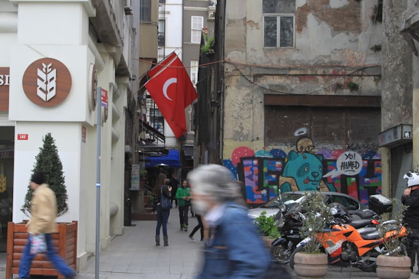 An urban street scene with a pedestrian walking by and a Turkish flag hanging prominently. The building on the right features a colorful graffiti mural with the words 'Yo! Ahmed' and a cartoonish character. Several motorcycles are parked in front of the mural. The left side of the image shows a storefront with a sign featuring a wheat symbol.