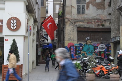 An urban street scene with a pedestrian walking by and a Turkish flag hanging prominently. The building on the right features a colorful graffiti mural with the words 'Yo! Ahmed' and a cartoonish character. Several motorcycles are parked in front of the mural. The left side of the image shows a storefront with a sign featuring a wheat symbol.