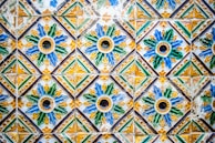 A vibrant and intricate pattern of ceramic tiles, each tile featuring a floral design in the center. The tiles are predominantly blue, green, yellow, and white, with each floral motif encircled by geometric shapes. The tiles show signs of aging and wear, adding character to the design.