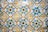 A vibrant and intricate pattern of ceramic tiles, each tile featuring a floral design in the center. The tiles are predominantly blue, green, yellow, and white, with each floral motif encircled by geometric shapes. The tiles show signs of aging and wear, adding character to the design.