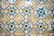 A vibrant and intricate pattern of ceramic tiles, each tile featuring a floral design in the center. The tiles are predominantly blue, green, yellow, and white, with each floral motif encircled by geometric shapes. The tiles show signs of aging and wear, adding character to the design.