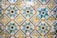 A vibrant and intricate pattern of ceramic tiles, each tile featuring a floral design in the center. The tiles are predominantly blue, green, yellow, and white, with each floral motif encircled by geometric shapes. The tiles show signs of aging and wear, adding character to the design.