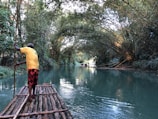 A man stands on a wooden raft navigating through a serene, narrow river flanked by lush greenery. The water is calm and reflects the surrounding foliage, creating a peaceful atmosphere. In the distance, another raft can be seen with people seated, enjoying the tranquil environment created by the canopy of trees arching overhead.