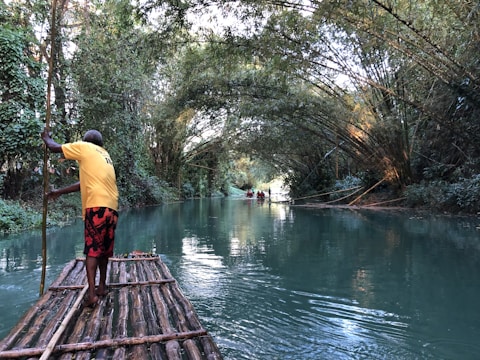 A man stands on a wooden raft navigating through a serene, narrow river flanked by lush greenery. The water is calm and reflects the surrounding foliage, creating a peaceful atmosphere. In the distance, another raft can be seen with people seated, enjoying the tranquil environment created by the canopy of trees arching overhead.