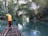 A man stands on a wooden raft navigating through a serene, narrow river flanked by lush greenery. The water is calm and reflects the surrounding foliage, creating a peaceful atmosphere. In the distance, another raft can be seen with people seated, enjoying the tranquil environment created by the canopy of trees arching overhead.