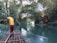 A man stands on a wooden raft navigating through a serene, narrow river flanked by lush greenery. The water is calm and reflects the surrounding foliage, creating a peaceful atmosphere. In the distance, another raft can be seen with people seated, enjoying the tranquil environment created by the canopy of trees arching overhead.