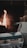 A cozy fireplace setting featuring a burning fire in the background, casting a warm glow. There is a woven blanket with a geometric pattern in shades of green, beige, and white, labeled 'sackcloth + ashes'. Next to it is a canvas basket filled with firewood.