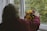 An elderly person with grey hair is seen from behind, wearing a maroon sweater, placing a colorful bouquet of flowers on a windowsill. The window is open, revealing a blurred view of green foliage outside.