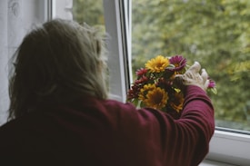 An elderly person with grey hair is seen from behind, wearing a maroon sweater, placing a colorful bouquet of flowers on a windowsill. The window is open, revealing a blurred view of green foliage outside.