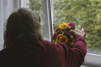 An elderly person with grey hair is seen from behind, wearing a maroon sweater, placing a colorful bouquet of flowers on a windowsill. The window is open, revealing a blurred view of green foliage outside.