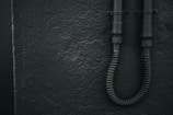 A textured wall with a rough finish is shown on the left. Two black, industrial-looking pipes are affixed to the wall on the right, with a flexible conduit at the bottom.