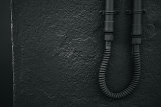 A textured wall with a rough finish is shown on the left. Two black, industrial-looking pipes are affixed to the wall on the right, with a flexible conduit at the bottom.