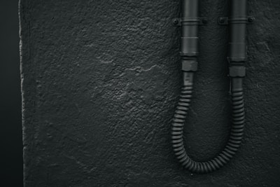 A textured wall with a rough finish is shown on the left. Two black, industrial-looking pipes are affixed to the wall on the right, with a flexible conduit at the bottom.