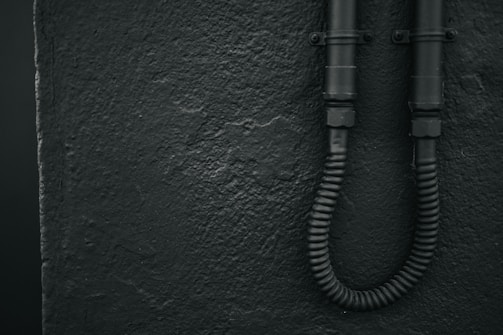 A textured wall with a rough finish is shown on the left. Two black, industrial-looking pipes are affixed to the wall on the right, with a flexible conduit at the bottom.