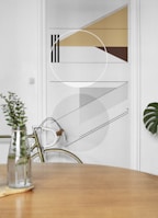 A minimalist interior featuring a geometric wall design with triangles, circles, and lines in shades of brown, gray, and beige. In the foreground, a wooden table with a glass vase containing green foliage. A bicycle with a white handlebar is partially visible, leaning against the wall.