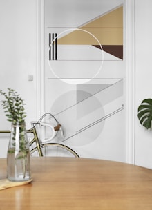 A minimalist interior featuring a geometric wall design with triangles, circles, and lines in shades of brown, gray, and beige. In the foreground, a wooden table with a glass vase containing green foliage. A bicycle with a white handlebar is partially visible, leaning against the wall.