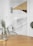 A minimalist interior featuring a geometric wall design with triangles, circles, and lines in shades of brown, gray, and beige. In the foreground, a wooden table with a glass vase containing green foliage. A bicycle with a white handlebar is partially visible, leaning against the wall.