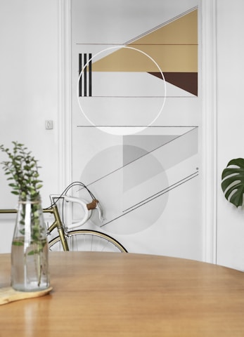 A minimalist interior featuring a geometric wall design with triangles, circles, and lines in shades of brown, gray, and beige. In the foreground, a wooden table with a glass vase containing green foliage. A bicycle with a white handlebar is partially visible, leaning against the wall.