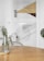 A minimalist interior featuring a geometric wall design with triangles, circles, and lines in shades of brown, gray, and beige. In the foreground, a wooden table with a glass vase containing green foliage. A bicycle with a white handlebar is partially visible, leaning against the wall.
