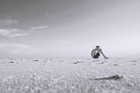 A solitary person sits on a vast, barren landscape under a wide, open sky. The scene is minimalistic, with the expanse of the sand appearing almost monochrome. The person is seated, knees drawn to their chest, gazing into the distance, creating a sense of solitude and introspection.