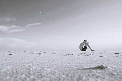 A solitary person sits on a vast, barren landscape under a wide, open sky. The scene is minimalistic, with the expanse of the sand appearing almost monochrome. The person is seated, knees drawn to their chest, gazing into the distance, creating a sense of solitude and introspection.